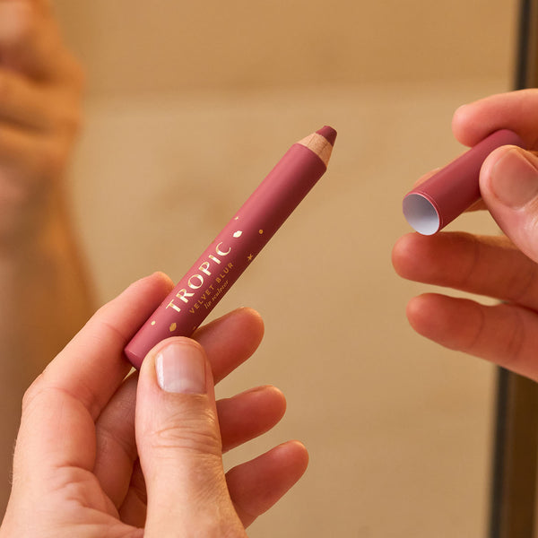 Close-up of a person holding Tropic’s Velvet Blur lip crayon from The Enhancing Lip Service collection, with the cap removed to reveal the soft, creamy lip colour.
