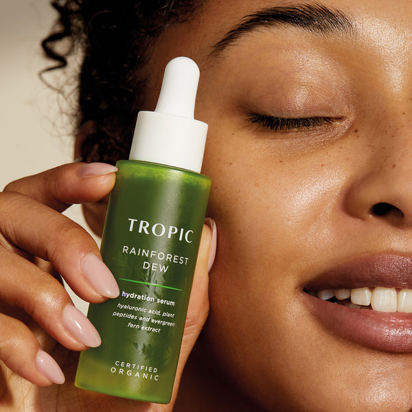 A close-up of a person with light to medium skin tone, smiling with their eyes closed. They are holding a green bottle labelled