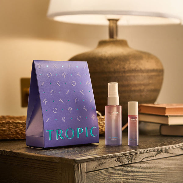 The Pillow Service Aromatherapy Sleep Collection by Tropic displayed on a bedside table, including a purple gift box, a pillow mist spray, and a rollerball, all designed to help promote relaxation and restful sleep.
