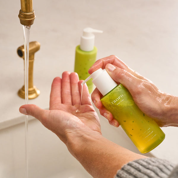 Person using Tropic’s Dessert Lime and Ginger Hand Wash, pumping the product into their hand under running water from a brass tap. A second bottle of the same product is visible in the background on the sink.
