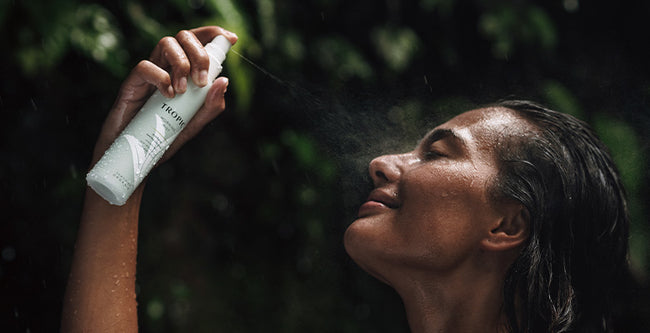 A woman with wet hair sprays a Tropic Morning mist onto her face, her eyes closed in relaxation. The mist is visible in the air, and the background features lush greenery, evoking a refreshing and natural skincare experience.