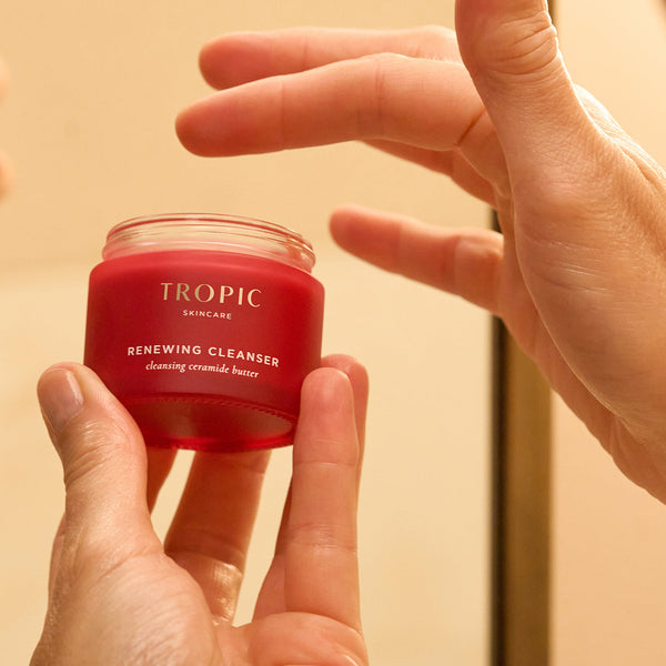 Close-up of hands holding Tropic Skincare Renewing Cleanser jar, with a fingertip about to scoop out the cleansing ceramide butter.