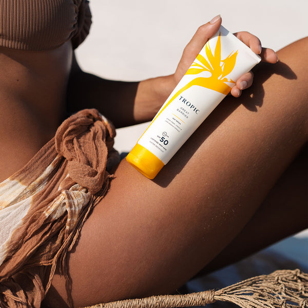 Model holding Tropic Great Barrier Sun Lotion SPF 50 while sitting on the beach, showing sun protection skincare in a summer setting.