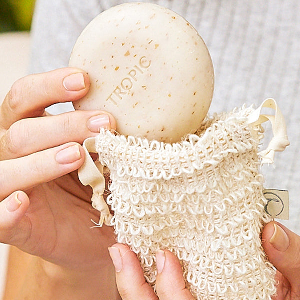 The image shows a person holding a Tropic Cleansing Pebble and a Body Pebble embossed with the brand name