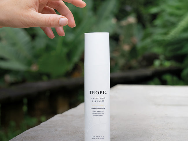 Hand reaching for Tropic Smoothing Cleanser bottle placed outdoors on a stone surface, with lush green foliage in the blurred background.