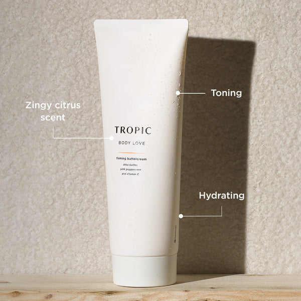 The image displays a tube of Tropic Body Love Firming Buttercream standing against a textured wall with sunlight casting a soft shadow. Key features of the product are labeled:
Zingy citrus scent, providing a refreshing, uplifting aroma.
Toning, aimed at improving skin firmness and elasticity.
Hydrating, formulated with nourishing ingredients like shea butter, pink peppercorn, and vitamin E to deeply moisturize the skin.