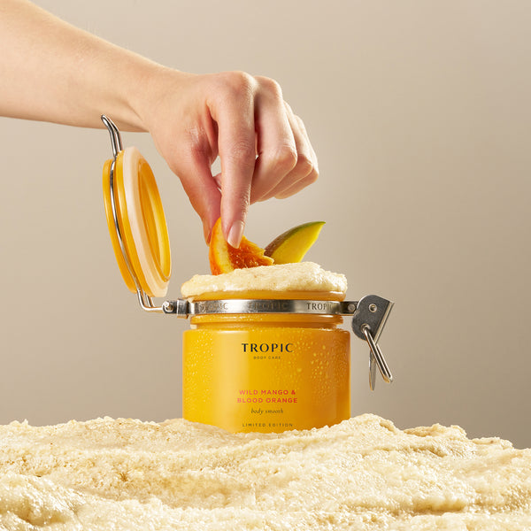 Hand placing a blood orange slice into an open jar of Tropic Wild Mango & Blood Orange Body Smooth, showcasing the whipped texture and fruity inspiration.