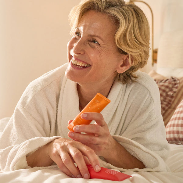 Smiling woman in a white robe sitting on a bed holding Tropic Skincare Spiced and Citrus body care products in orange and red tubes.