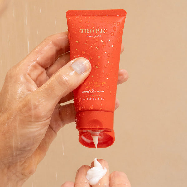 Close-up of Tropic Spiced Citrus & Clove Creamy Body Cleanser in a red limited-edition tube, with water droplets as it’s dispensed onto a hand under the shower.