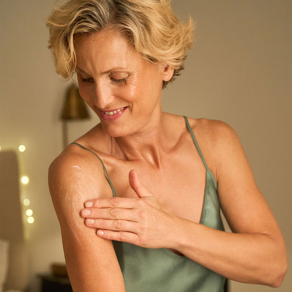 A smiling woman with short blonde hair applies lotion to her upper arm, wearing a green satin camisole, in a softly lit room.