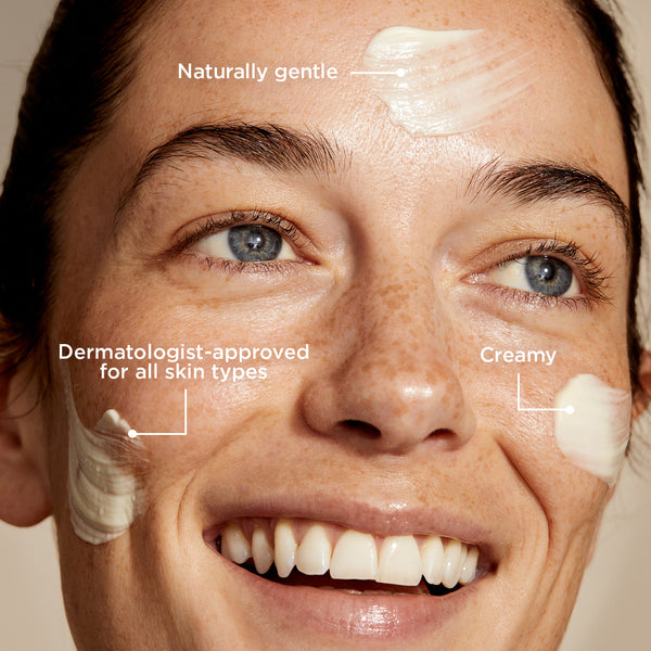 A close-up of a smiling person with light skin and blue eyes, with a few small dabs of smoothing cleanser applied to their face—specifically on the forehead and cheeks. Text annotations on the image highlight that the product is 