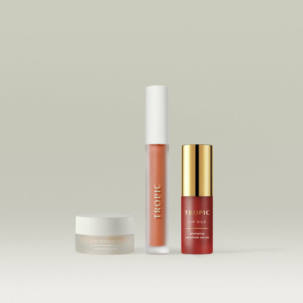 Tropic The Full Lip Service collection, featuring Lip Smooth Softening Scrub in a frosted jar, a Lip Stain in a burnt coral shade with a sleek white cap, and Lip Silk Plumping Ceramide Serum in a gold-capped bottle. A luxurious trio for nourished, hydrated, and beautifully tinted lips.