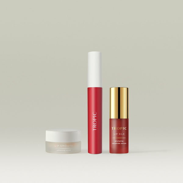 Tropic The Full Lip Service collection, featuring Lip Smooth Softening Scrub in a frosted jar, a Lip Stain in a vibrant red shade with a sleek white cap, and Lip Silk Plumping Ceramide Serum in a gold-capped bottle. A luxurious trio for nourished, hydrated, and beautifully tinted lips.
