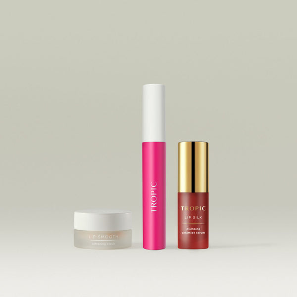 Tropic The Full Lip Service collection, featuring Lip Smooth Softening Scrub in a frosted jar, a Lip Stain in a vibrant pink shade with a sleek white cap, and Lip Silk Plumping Ceramide Serum in a gold-capped bottle. A luxurious trio for nourished, hydrated, and beautifully tinted lips.