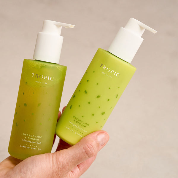 Close-up of a hand holding two Tropic Body Care products from the Desert Lime & Ginger Limited Edition range. One bottle is labelled “Energising Hand Wash” and the other “Enveloping Hand Cream,” both in bright green packaging with white pump dispensers.