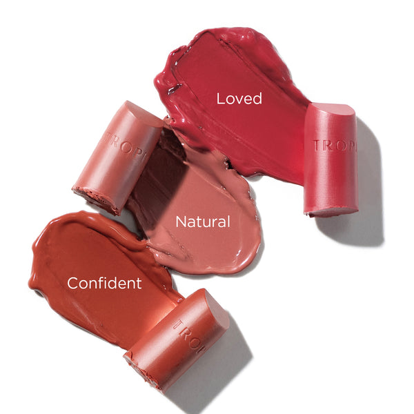 Three swatches of lipstick shades with broken lipstick bullets labelled 'Loved' (A delicate petal pink), 'Natural' (A sheer effortless netural), and 'Confident' (A soft burnt rouge). The brand name 'Tropic' is embossed on each lipstick bullet.