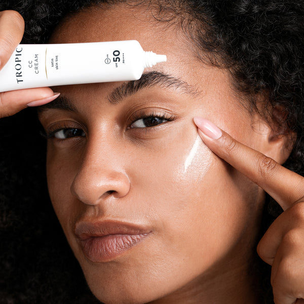 A close-up of a person applying Tropic CC cream with SPF 50 to their forehead and cheek. The individual has curly hair, medium-toned skin, and is gently pressing the product into their skin with their fingers, while holding the tube of cream in one hand.