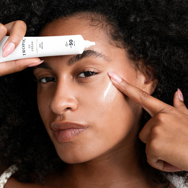 A close-up of a person applying Tropic CC cream with SPF 50 to their forehead and cheek. The individual has curly hair, medium-toned skin, and is gently pressing the product into their skin with their fingers, while holding the tube of cream in one hand.
