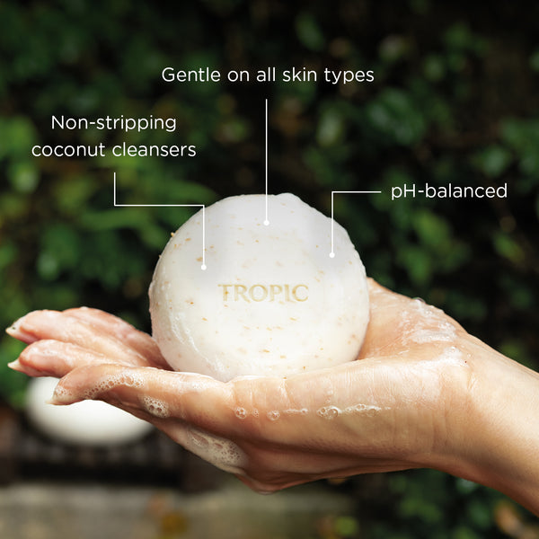 Hand holding a Tropic body pebble with text highlighting its benefits on a blurred green background; non-stripping coconut cleansers, pH-balanced and gentle on all skin types.