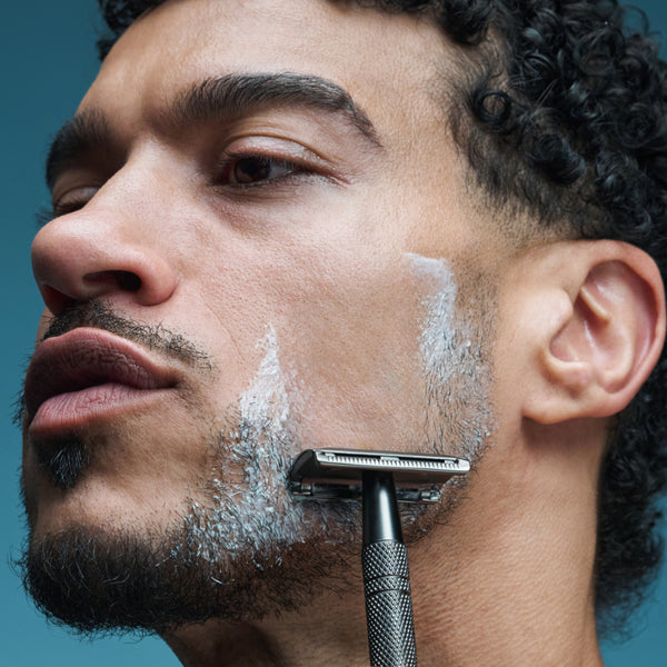 Close-up of model shaving with razor and Tropic Look Sharp shaving cream applied to face.
