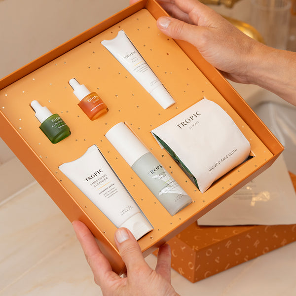 Hands holding an open orange gift box containing Tropic Skincare products, including mini serums, cleanser, moisturiser, toner, and a bamboo face cloth.