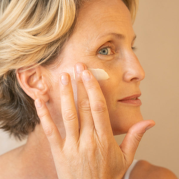 Close-up of a woman gently applying a small amount of product to her cheek with her fingertips, smiling softly.