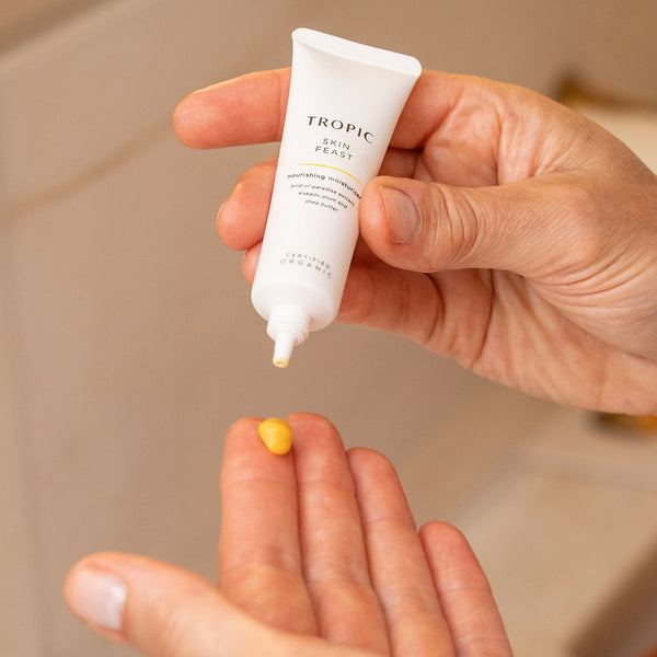 Hands dispensing a small amount of Tropic Skin Feast Nourishing Moisturiser from a white tube, with the golden-yellow cream placed onto a fingertip.