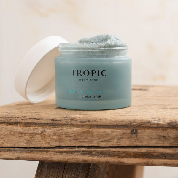 An open jar of Tropic Body Care Foot Smooth Enzymatic Scrub with a white lid resting to the side, placed on a rustic wooden surface.