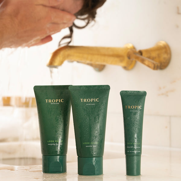 Tropic Skincare products on a bathroom counter with water droplets, featuring Look Fresh, Look Sharp, and Skin Shield in dark green tubes, with a person washing their face in the background.