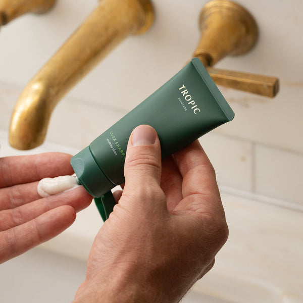 Hands squeezing Tropic Skincare’s green tube of Look Sharp shave cream, dispensing some of the product onto their fingers, with brass bathroom taps in the background.