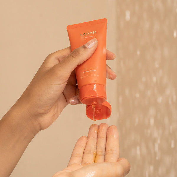 Hand squeezing Tropic Body Care Mango Grove gel body cleanser from an orange tube into the palm, with water droplets in the background.