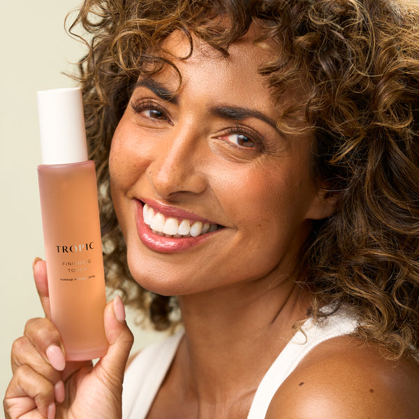 A smiling woman holding Tropic Finishing Touch setting spray, showcasing a radiant, natural glow.