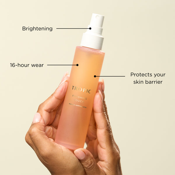 A hand holding Tropic Finishing Touch setting spray, highlighting brightening, 16-hour wear, and skin barrier protection.