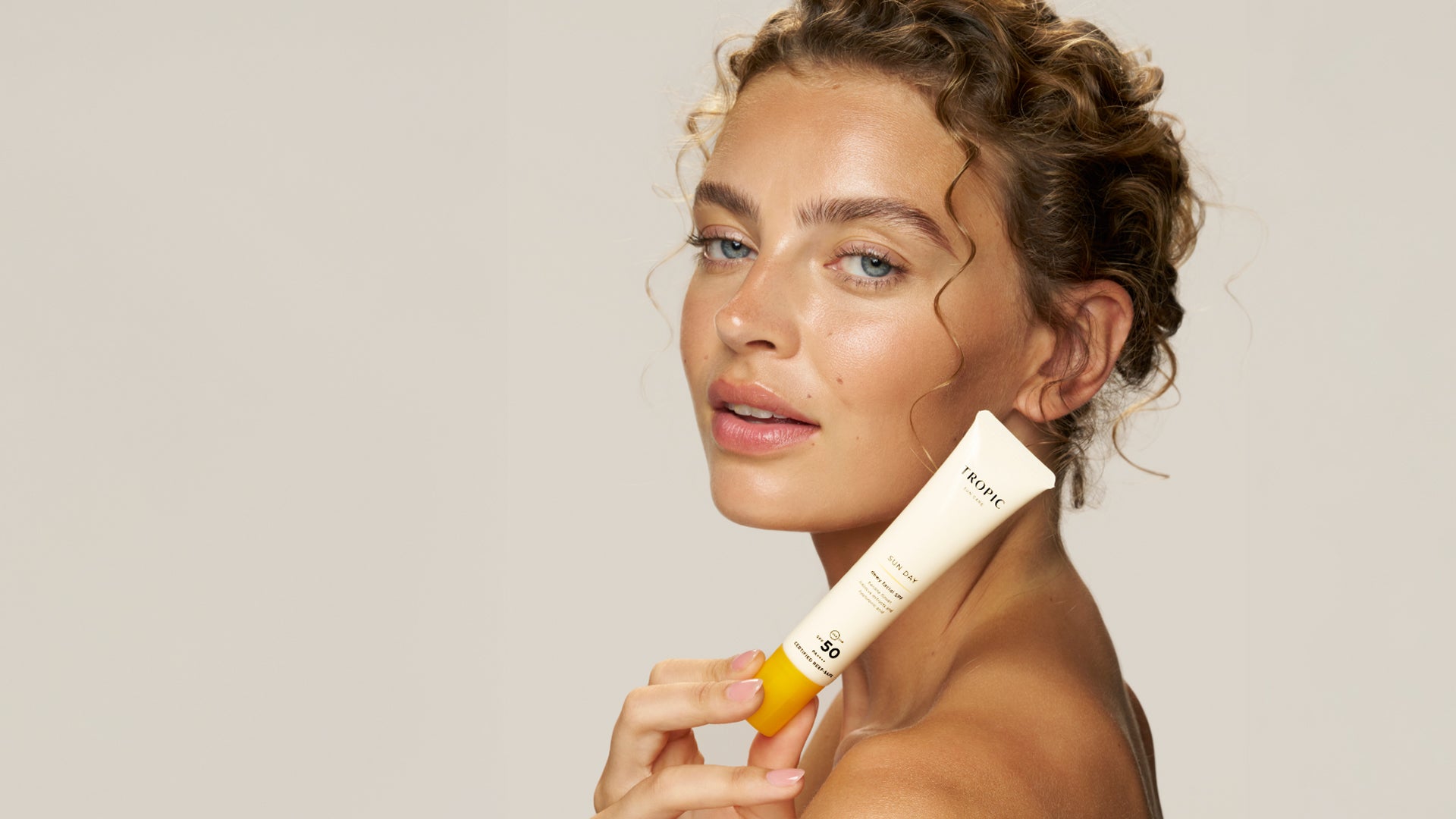 Woman holding a tube of Sunday SPF protection above her shoulder with a neutral background