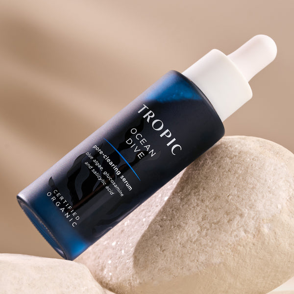 This image showcases a bottle of Ocean Dive, a pore-clearing serum. The dark blue packaging, featuring a dropper applicator, highlights its key ingredients: blue algae glucosamine and salicylic acid. The product is certified organic and is designed to refine pores, clear skin, and promote a smooth complexion. It is elegantly displayed on a natural stone surface, emphasising its eco-conscious and natural formulation.