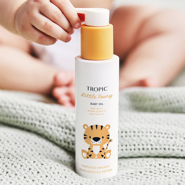 The image showcases a bottle of Tropic Little Roary Baby Oil, held by a baby with soft, cozy blankets in the background. The bottle features a cheerful tiger illustration, appealing to children and parents alike. The label highlights nourishing ingredients such as tiger grass, golden jojoba oil, and vitamin E, which help soothe and moisturize delicate baby skin. The product is marked as Certified Organic and Pediatrician Tested, emphasizing its safety and natural formulation.