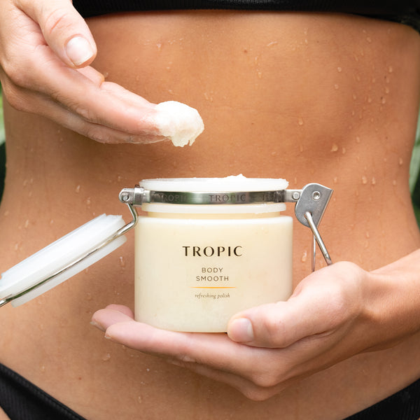 Model scoops product into their hands from Tropic Body Smooth refreshing polish jar. The tub has a silver clasp with Tropic written on it. The model holds the jar with the other hand against her stomach which has water droplets on.