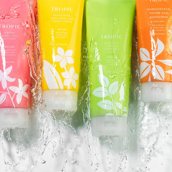 The image features a vibrant lineup of Tropic Body Washes, each in brightly colored packaging with botanical designs. The tubes are partially submerged in cascading water, emphasizing their refreshing and hydrating qualities. The visible scents include:

Jasmine, Neroli and Vetiver (pink).
Frangipani, Star Anise and Tiaré Flower (yellow).
Wild Mint, Lime and Pineapple (green).
Mangosteen, Thyme and Mandarin (orange).
Each body wash is pH-balanced, highlighting their gentle yet effective cleansing properties