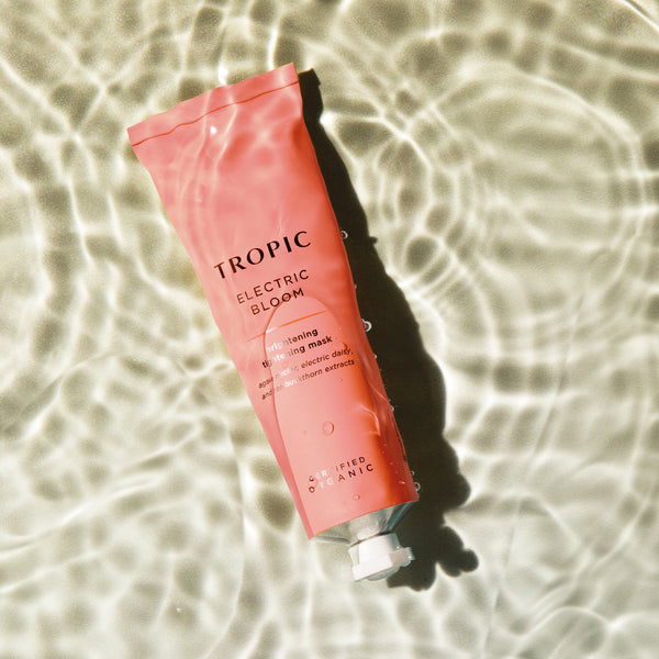 This image features a tube of Electric Bloom, a brightening tightening mask. The coral pink packaging highlights its vibrant and rejuvenating properties. Key ingredients include bagasse water, electric daisy, and sea buckthorn extracts. The product is certified organic, and the rippling water backdrop adds a refreshing and invigorating aesthetic.