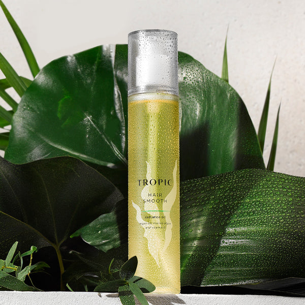 A sleek bottle of Tropic Hair Smooth Radiance Oil against a backdrop of fresh green leaves. The bottle is translucent, revealing a golden-yellow liquid inside, and it is adorned with water droplets to evoke a refreshing and natural feel. The label highlights key ingredients: argan oil, maracuja oil, and vitamin E, emphasizing its nourishing and smoothing benefits. The lush greenery complements the product’s natural and luxurious appeal.