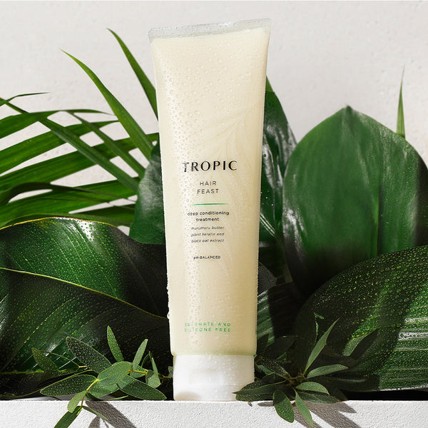 The image features a tube of Tropic Hair Feast Deep Conditioning Treatment positioned against a backdrop of vibrant green foliage. The tube is pale cream in color, with water droplets on its surface, evoking a sense of hydration and freshness. The label highlights key nourishing ingredients such as murumuru butter, plant keratin, and black oat extract. It is described as pH-balanced and sulphate and silicone free, emphasizing its gentle, moisturizing, and restorative properties for the hair.