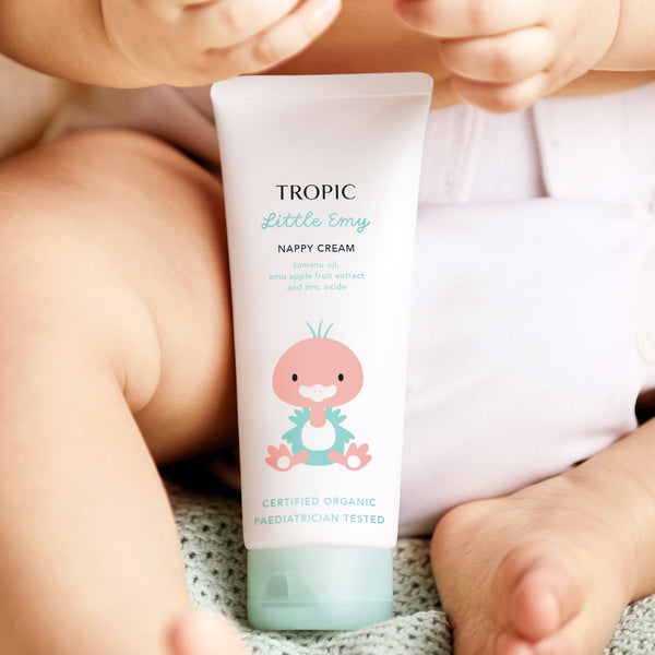 The image displays a tube of Tropic Little Emy Nappy Cream held by a baby, with their legs and diaper visible in the background. The packaging features a cute illustration of a pink bird, creating a playful and child-friendly design. The label highlights key ingredients such as tamanu oil, emu apple fruit extract, and zinc oxide, known for their soothing and protective properties. It is Certified Organic and Pediatrician Tested, emphasizing its safety and suitability for delicate baby skin.