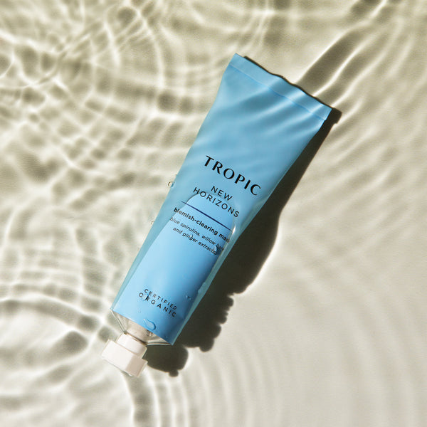 This image displays a tube of New Horizons, a blemish-clearing mask. The light blue packaging reflects its calming and clarifying properties. Key ingredients include blue spirulina, willow bark, and ginger extracts, formulated to target blemishes and promote clearer skin. The rippling water effect in the background emphasises its refreshing and purifying benefits. The product is certified organic, highlighting its natural and gentle approach to skincare.