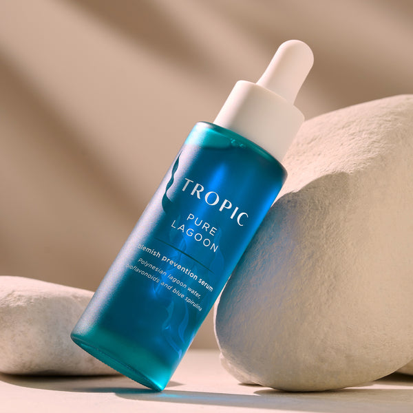 This image features a bottle of Pure Lagoon, a blemish prevention serum. The sleek blue packaging is paired with a dropper applicator, exuding a fresh and calming aesthetic. Key ingredients include Polynesian lagoon water, bioflavonoids, and blue spirulina, designed to help prevent blemishes while soothing and balancing the skin. The bottle is displayed against smooth, natural stones, enhancing its clean and pure skincare focus.