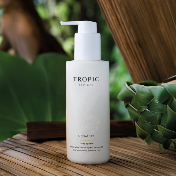 Tropic Body Care Signature hand wash is shown in a white bottle with white dispensing pump. There is a faint floral design on the bottle. The product contains Australian lemon myrtle, bergamot and eucalyptus essential oils. Its displayed on a wooden surface against a leafy background.