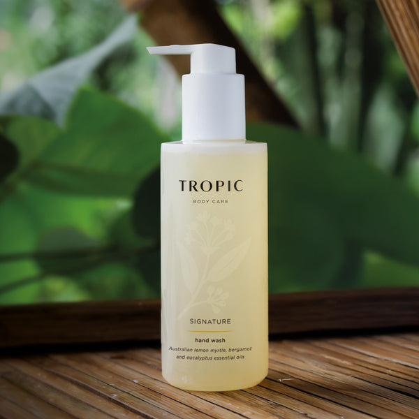 Tropic Body Care Signature Hand wash has a white dispensing pump and amber colour bottle with faint floral design. The product contains Australian lemon myrtle, bergamot and eucalyptus essential oils. The product is on a wooden surface with a leafy background.
