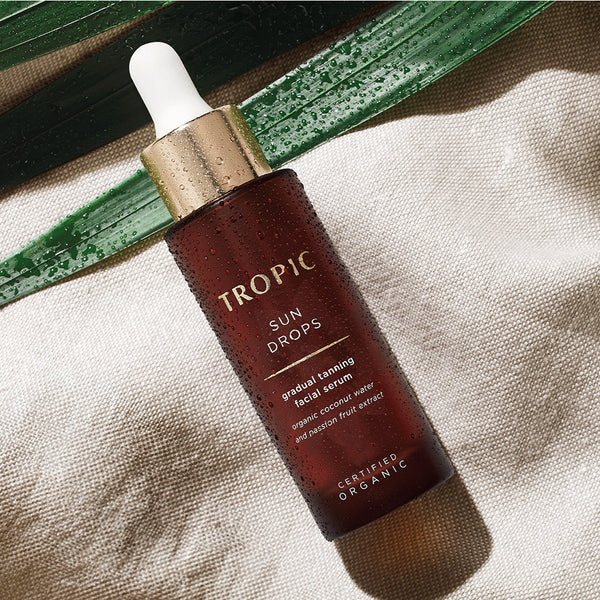 This image features a bottle of Sun Drops, a gradual tanning facial serum, resting on a textured fabric surface. The bottle is adorned with water droplets, enhancing its fresh, hydrating appeal. It is accompanied by a natural element, likely an aloe leaf, creating a refreshing and organic aesthetic. The product highlights include organic coconut water and passion fruit extract, emphasising its certified organic formulation.