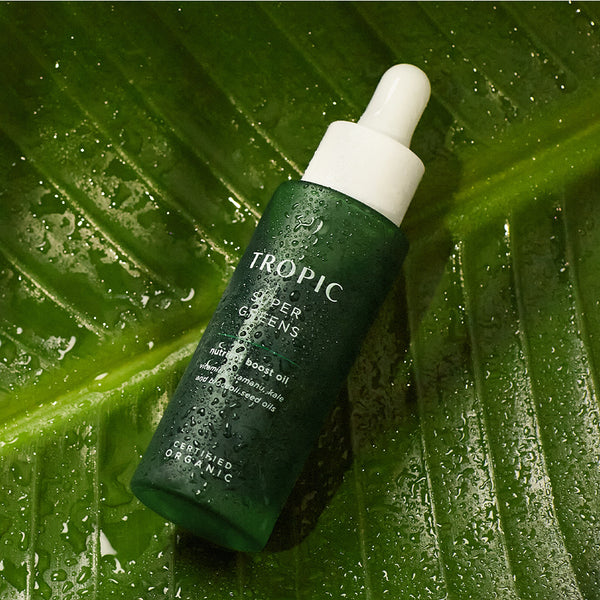 This image showcases a bottle of Tropic Super Greens, a nutrient boost oil. The deep green packaging is accentuated with water droplets, enhancing its fresh and natural appeal. Key ingredients include tamanu, kale, and broccoli seed oils, designed to deeply nourish, repair, and revitalize the skin. The bottle is elegantly placed on a large, glossy green leaf, reinforcing its organic and plant-based formulation.