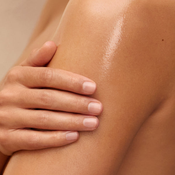 The image shows a close-up of a person’s shoulder and arm, with their hand gently resting on the skin. The skin appears smooth, hydrated, and glowing, suggesting the application of a rich, nourishing product like Tropic Whipped Body Velvet. The soft lighting highlights the healthy, radiant finish, evoking a sense of luxury and deep hydration.