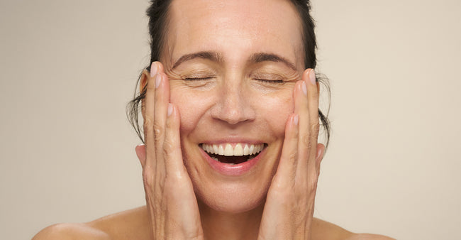 Smiling woman with eyes closed gently pressing her hands to her face, skin glowing against a neutral background.